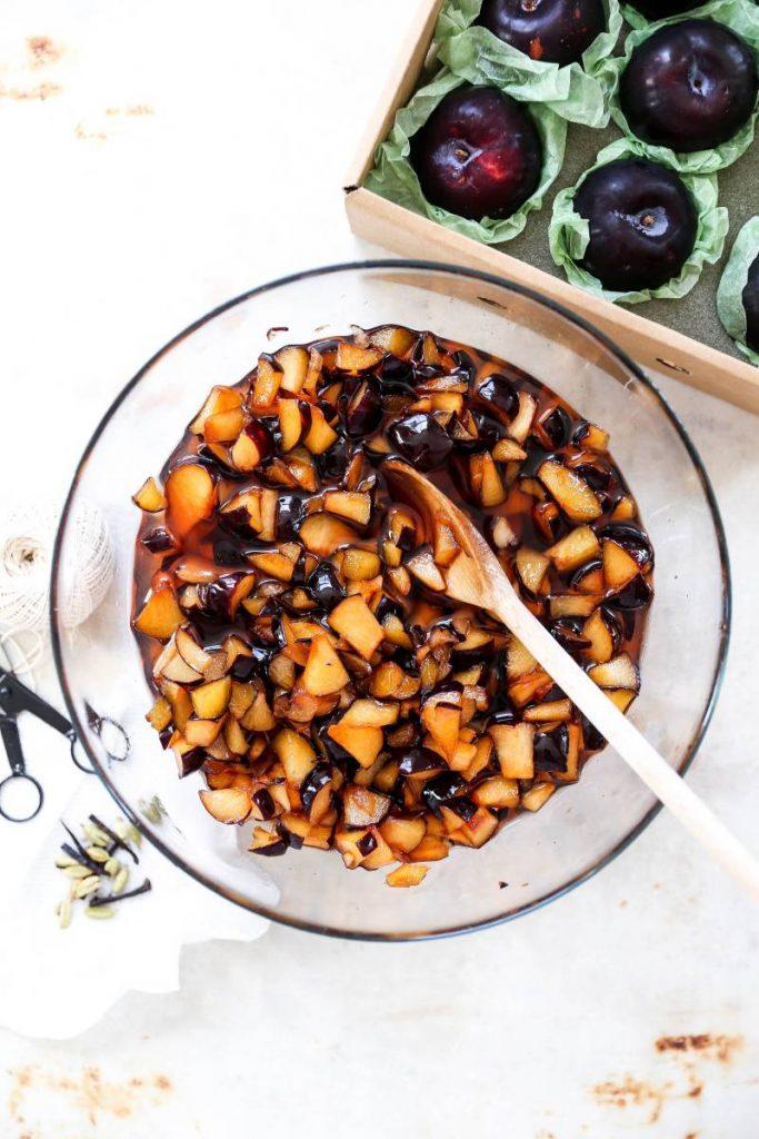 A photo of plum jam with a bowl of chopped plums next to a box of fresh, whole plums